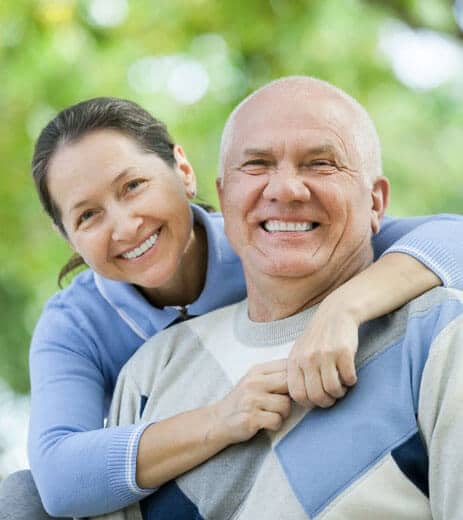Smiling middle-aged couple embracing outdoors with green foliage background.