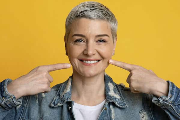 Smiling woman with short gray hair pointing at teeth against yellow background, dental promotion photo