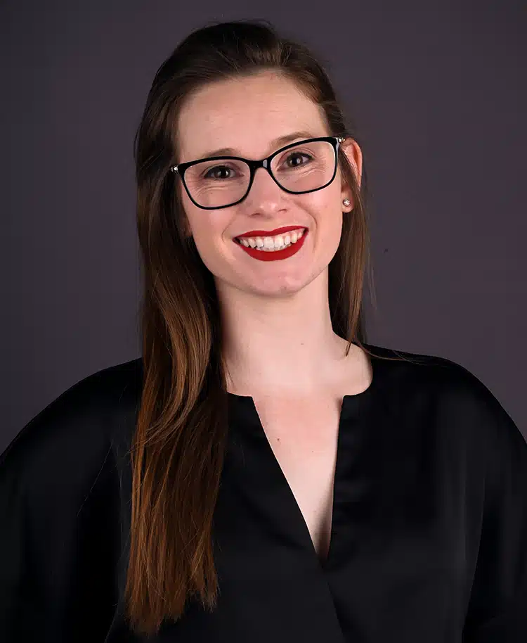Woman with glasses and red lipstick smiling at camera in professional headshot against gray background.