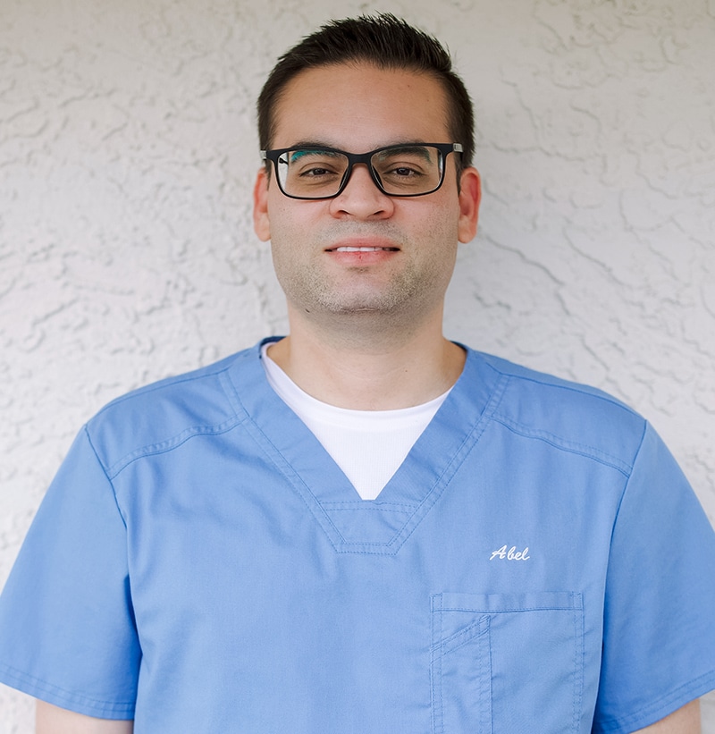 Male dental professional wearing blue scrubs and glasses against white background.